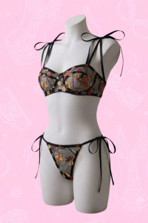 Fotografía de estudio de un maniquí blanco de medio cuerpo sobre un fondo rosa claro con patrones lineales tenues de lencería. El maniquí viste un conjunto de lencería de dos piezas confeccionado en malla negra transparente con bordados florales detallados en tonos amarillos, naranjas y rojos. El sostén y la braga estilo tanga se ajustan mediante cintas de satén negro muy delgadas que forman delicados lazos en los hombros y a los costados de la cadera.