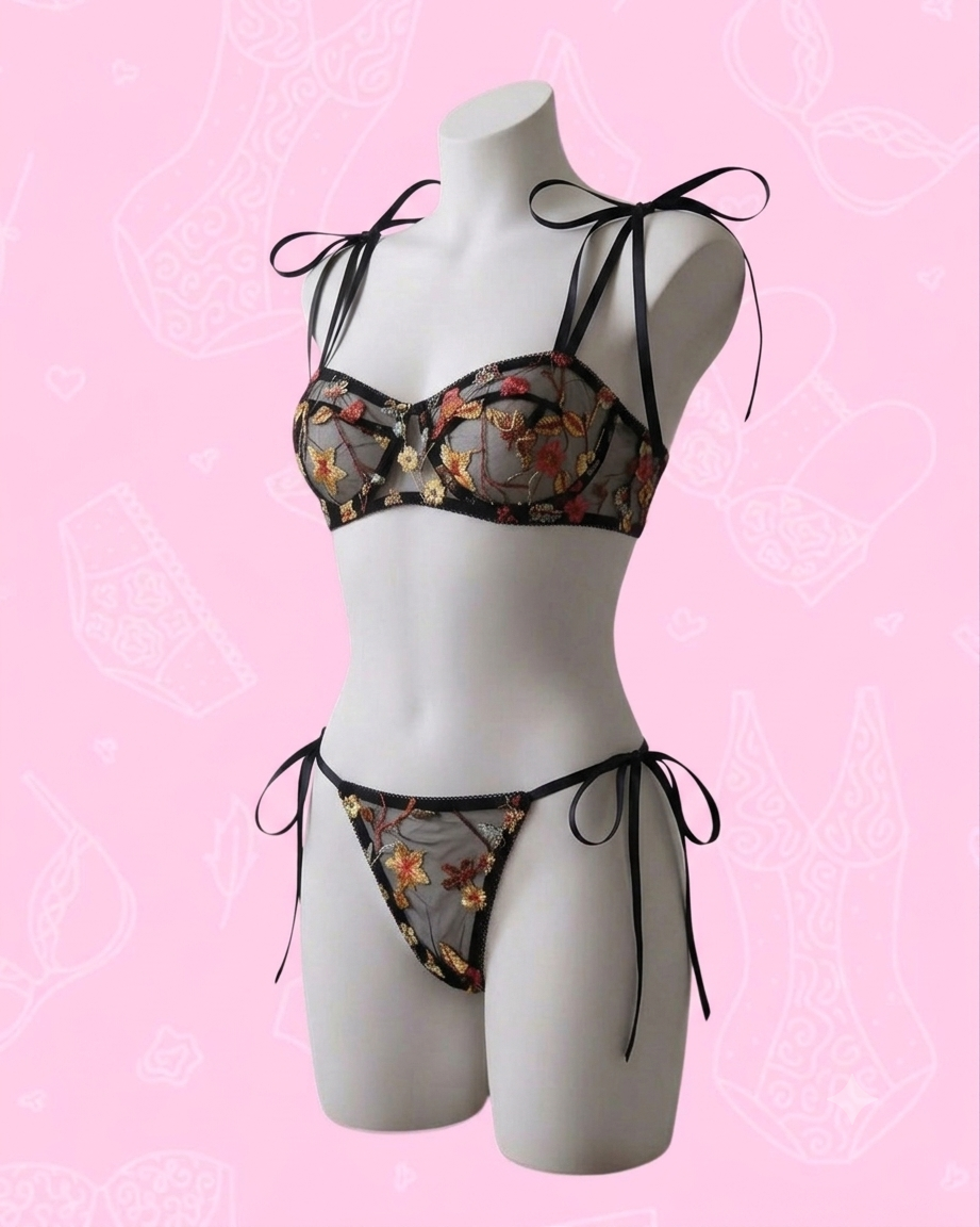 Fotografía de estudio de un maniquí blanco de medio cuerpo sobre un fondo rosa claro con patrones lineales tenues de lencería. El maniquí viste un conjunto de lencería de dos piezas confeccionado en malla negra transparente con bordados florales detallados en tonos amarillos, naranjas y rojos. El sostén y la braga estilo tanga se ajustan mediante cintas de satén negro muy delgadas que forman delicados lazos en los hombros y a los costados de la cadera.