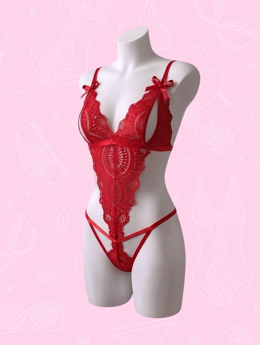 Un maniquí de torso femenino blanco, de perfil y de tres cuartos, muestra un body de lencería de encaje rojo sobre un fondo rosa suave decorado con contornos de lencería de color rosa más claro. El body cuenta con un sujetador estilo bralette de encaje con copas abiertas y lazos de satén rojo en las correas de los hombros, un panel central de encaje intrincado y una braga tanga de tiras en la parte inferior. El encaje tiene patrones florales y de medallón detallados.