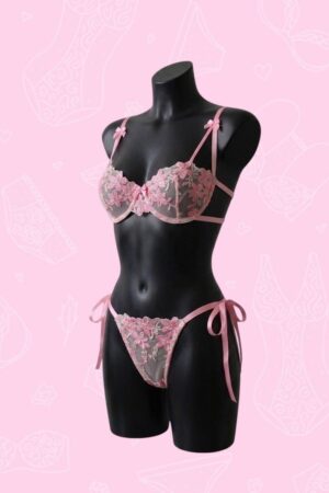 Conjunto de lencería rosa pastel con bordado floral de encaje en un maniquí blanco sobre fondo rosa decorado.